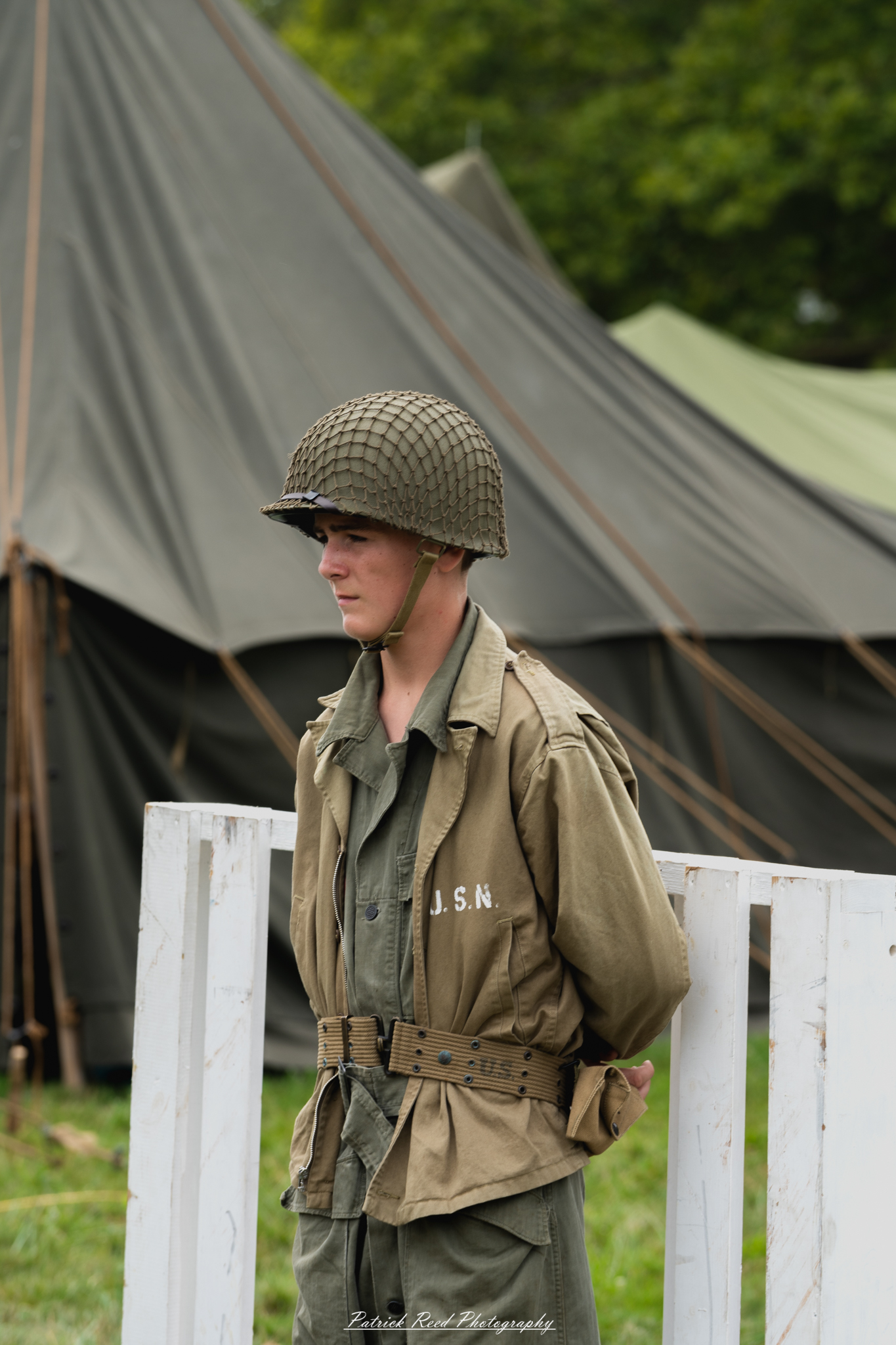 A young soldier standing alone, dressed in uniform with a thoughtful expression. His posture reflects a mix of determination and introspection, capturing the complexities of youth and responsibility in a military context.